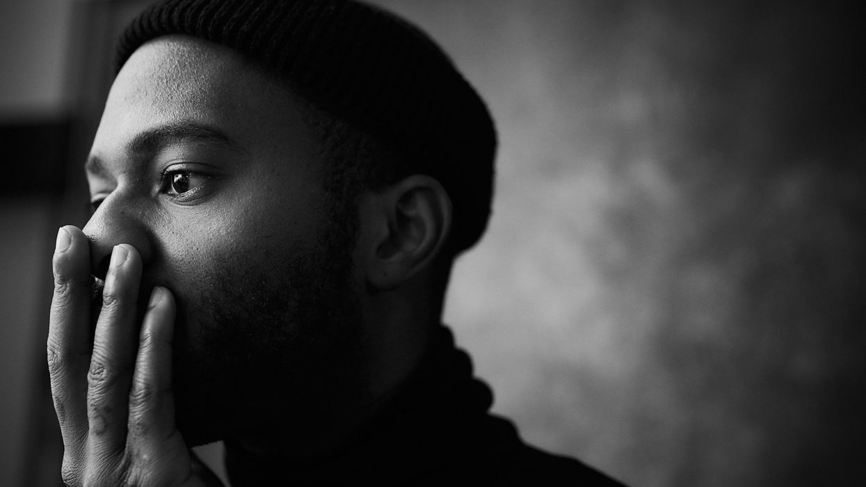 A black and white photo of a young man holding his hand to his face in shock, he wears a beanie.