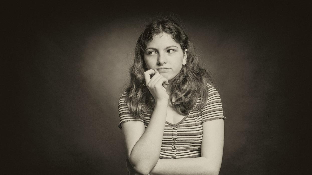 A black and white photo of a young woman in deep thought standing in front of a black background.