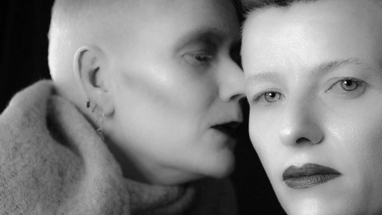 A black and white photo of two women. They have very short haircuts. One woman whispers into the other's ear.