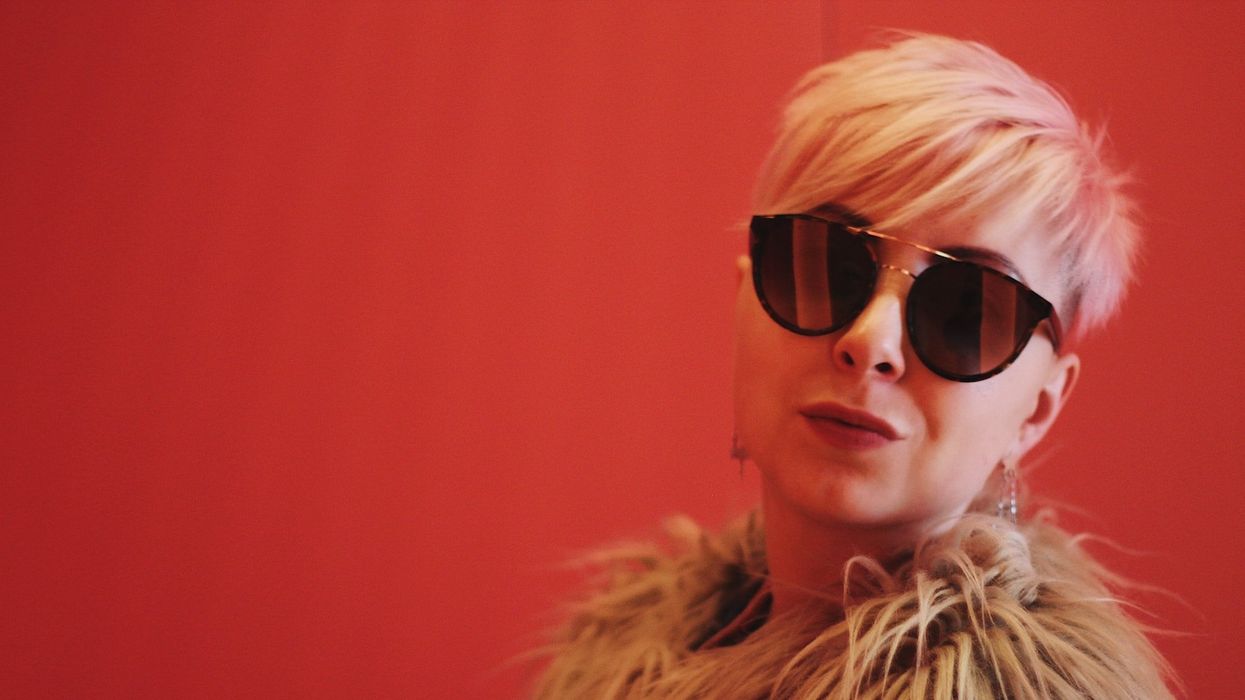 A blonde woman with sunglasses poses into the camera against a red background