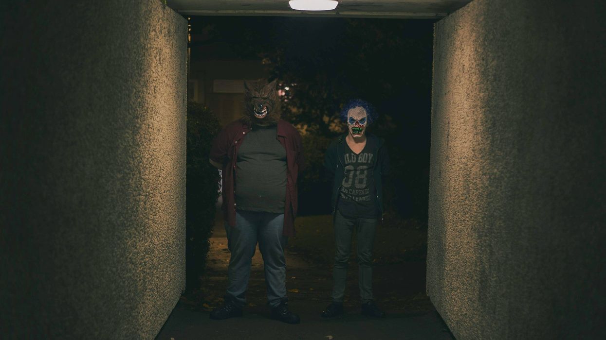 A creepy photo of two people standing eerily in a dark corridor, staring forward. One person wears a werewolf mask, the other a deranged clown mask.