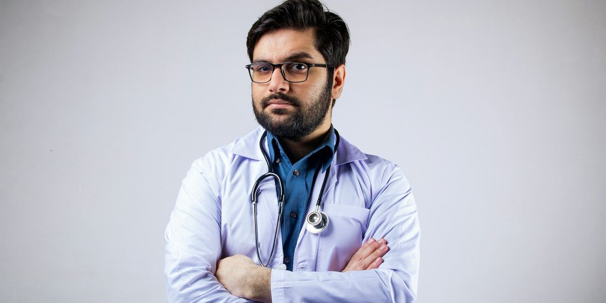 A doctor looks into the camera with an eyebrow raised. He has a stethoscope around his neck and his arms folded. 