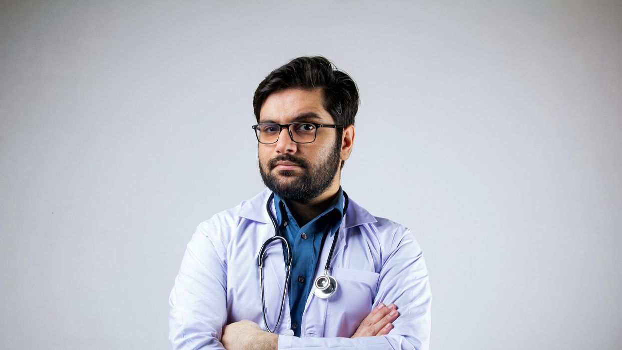 A doctor looks into the camera with an eyebrow raised. He has a stethoscope around his neck and his arms folded.
