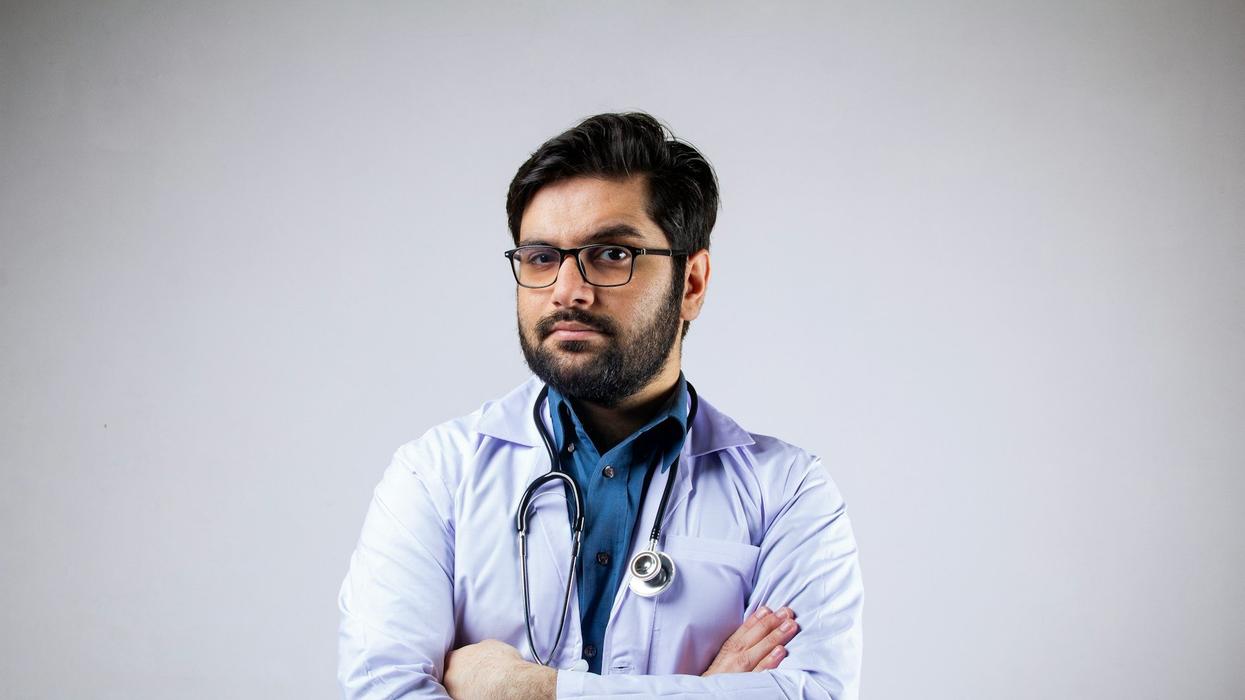 A doctor looks into the camera with an eyebrow raised. He has a stethoscope around his neck and his arms folded.