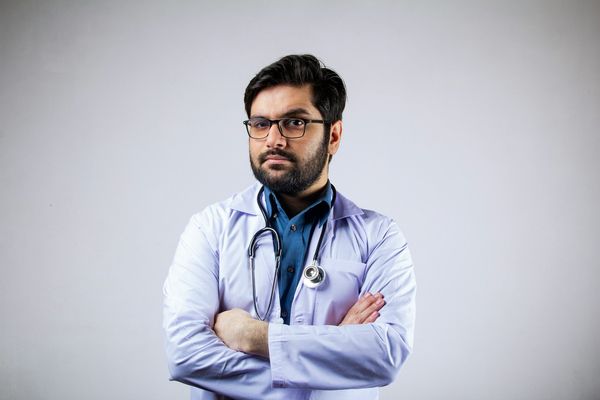 A doctor looks into the camera with an eyebrow raised. He has a stethoscope around his neck and his arms folded.