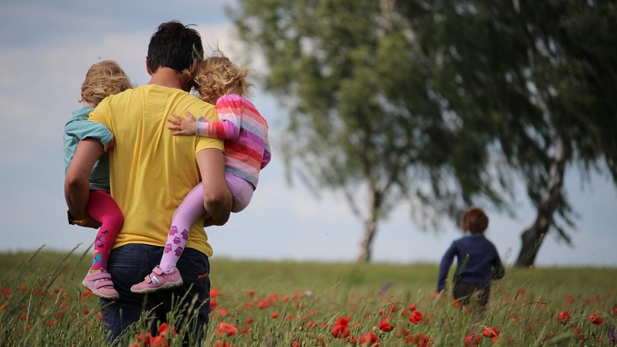 A father carrying two toddlers while observing a boy running ahead of him in a field