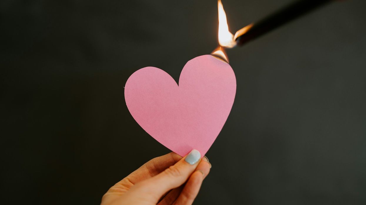 A female hand holds a pink paper heart that she is also setting on fire.