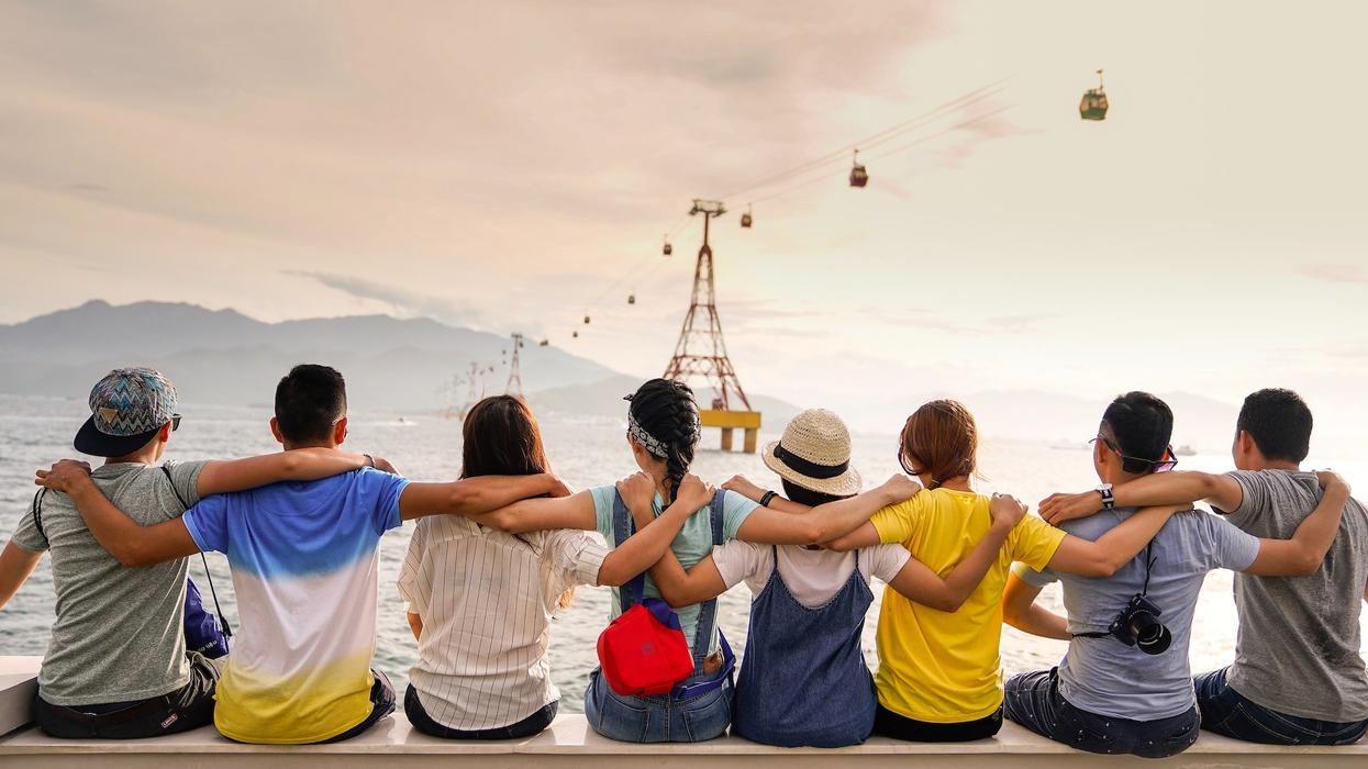 A group of friends sit with their arms around one another's shoulders while looking out into the sea