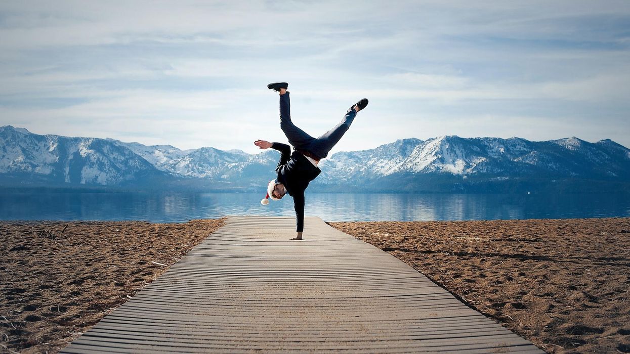 A guy in a Santa hat does a one armed hand stand with a mountain view behind him