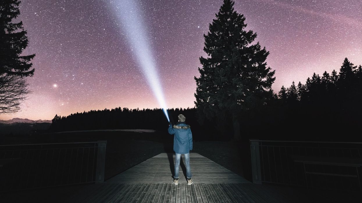 A guy stands on a dock, he holds up a flashlight into the night sky, stargazing
