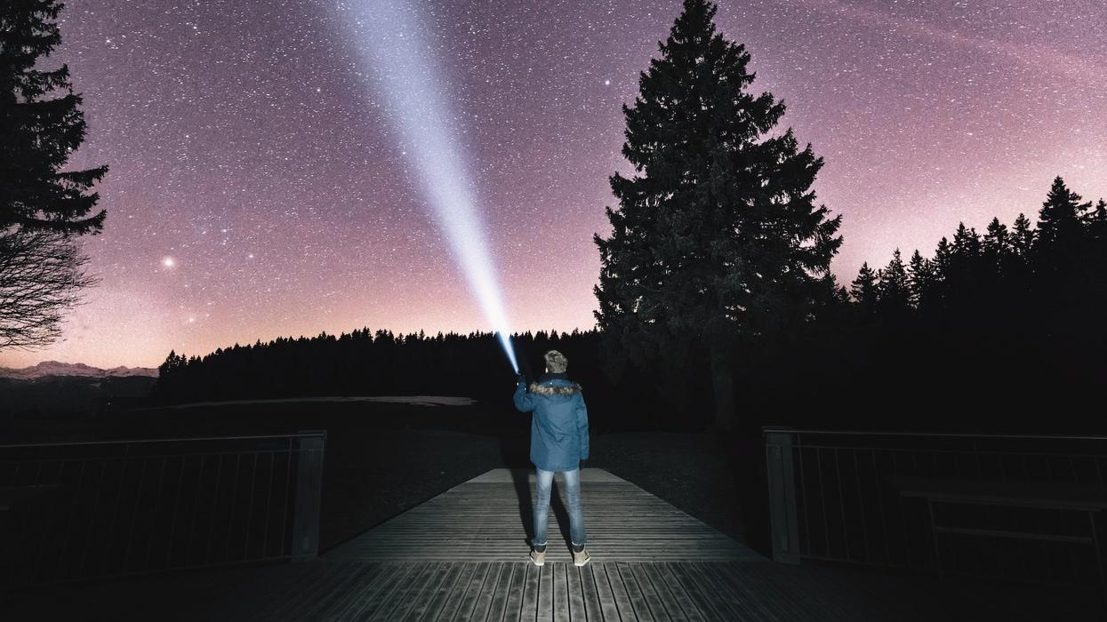 A guy stands on a dock, he holds up a flashlight into the night sky, stargazing