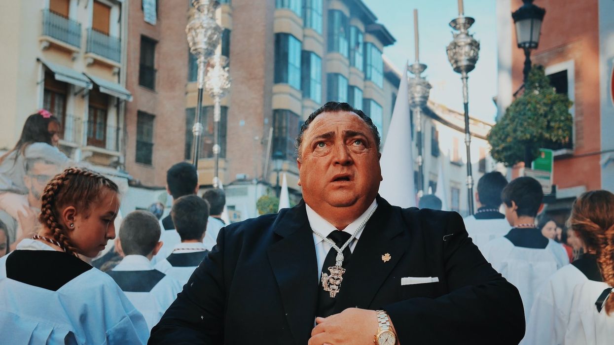 A man in a suit surrounded by altar boys and girls looks to the sky with a nervous facial expression