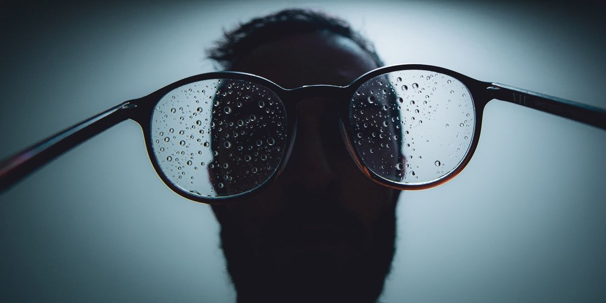 A man in shadow holds water spotted eyeglasses backwards 