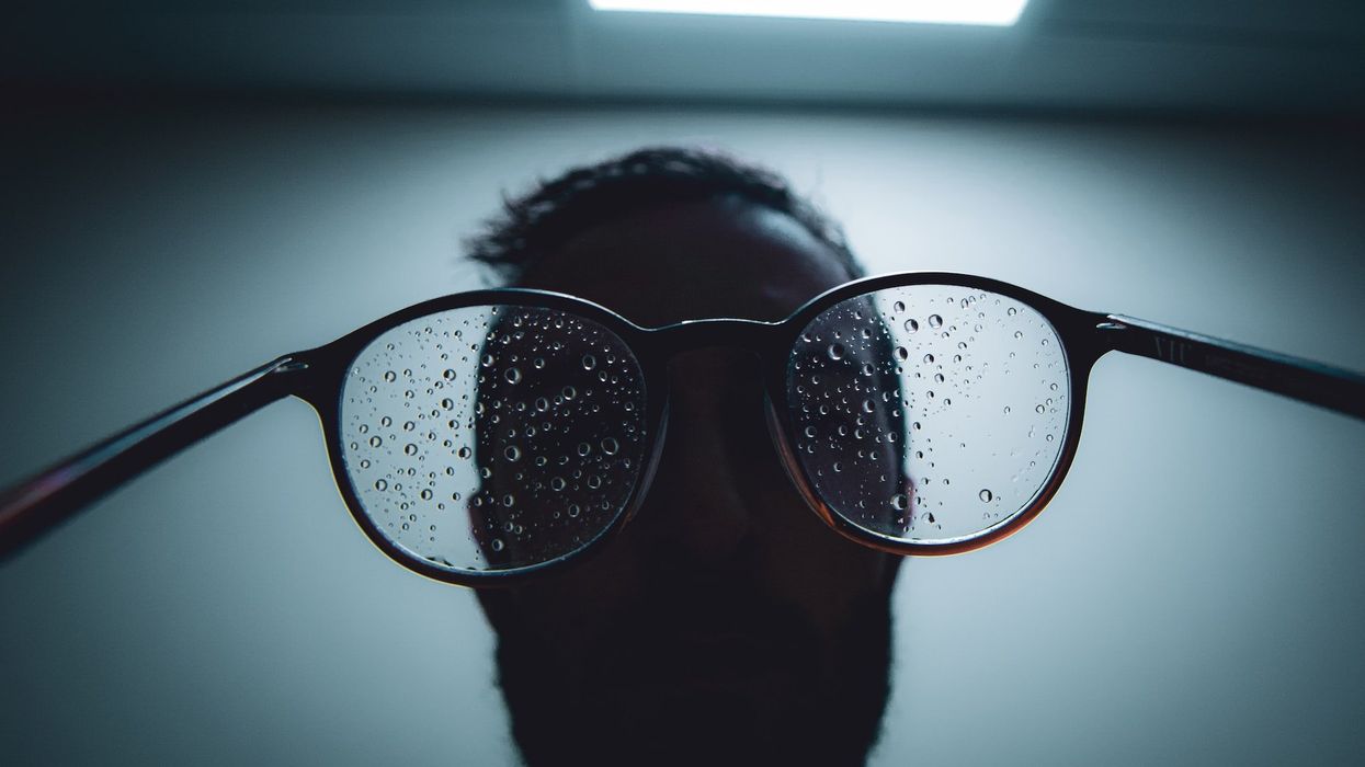 A man in shadow holds water spotted eyeglasses backwards