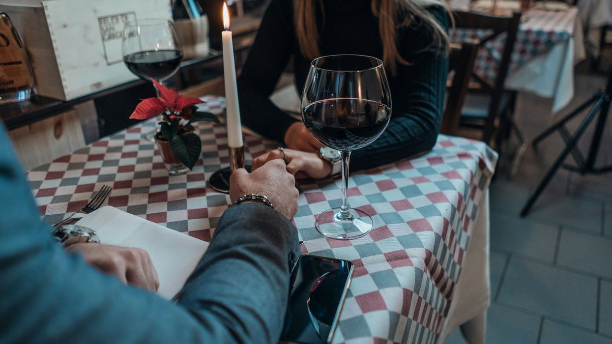 A mid-shot of a couple holding hands on a date. They are drinking red wine and a long stem candle is lit between. them.