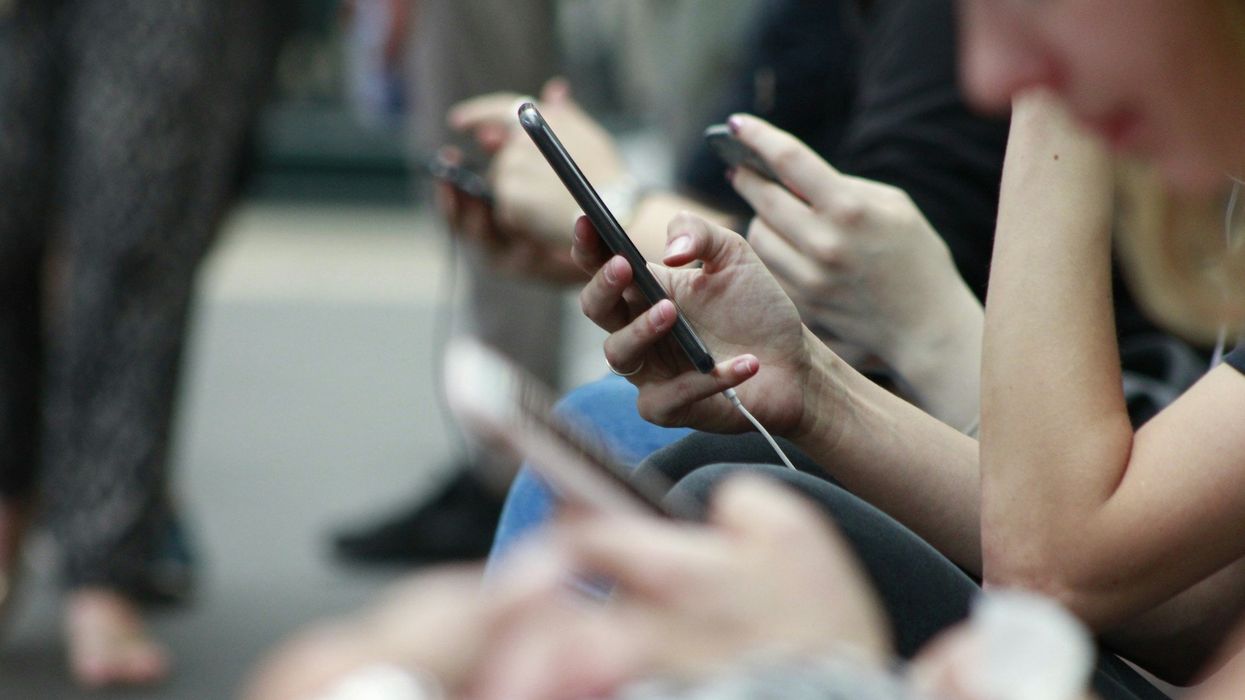 A mid-shot of a line of people in the park, looking into their cellphones. One phone and hand in particular is clear while the others are blurred.