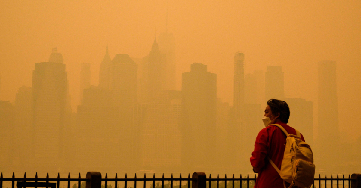A person with dark hear wearing a red jacket and light-colored backpack stands in front of a New York City skyline shrouded in orange smoke from the wildfires