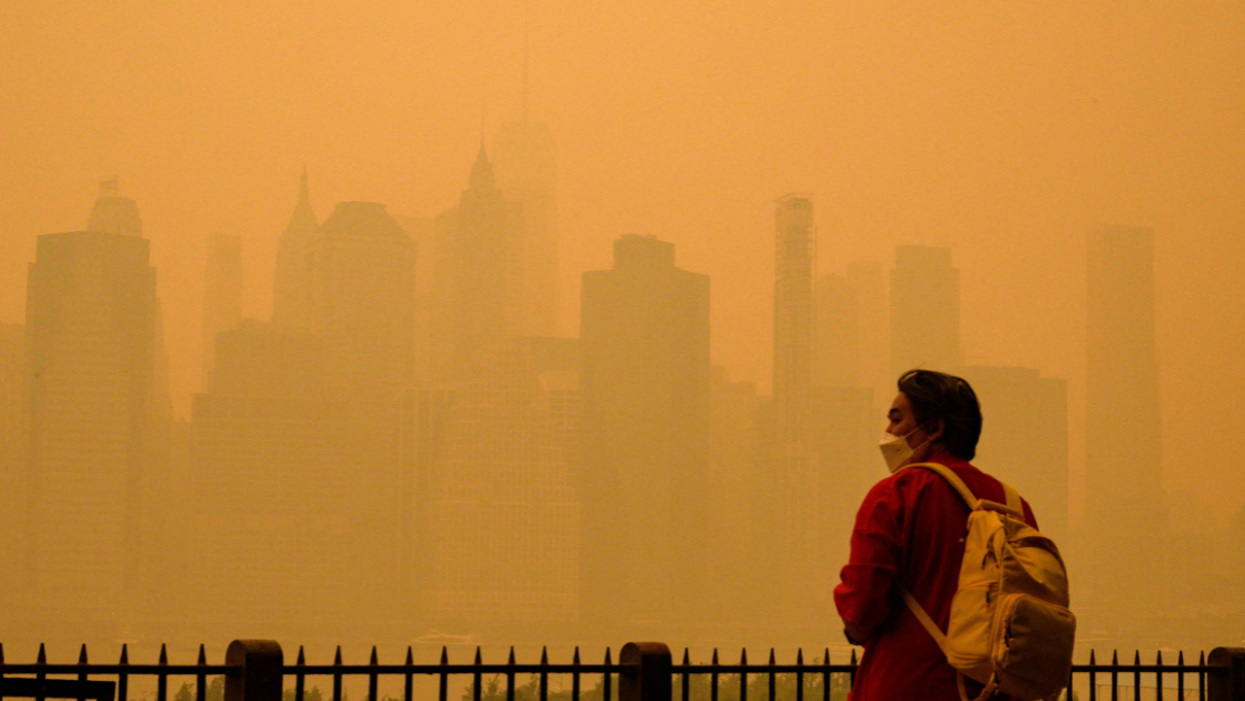 A person with dark hear wearing a red jacket and light-colored backpack stands in front of a New York City skyline shrouded in orange smoke from the wildfires