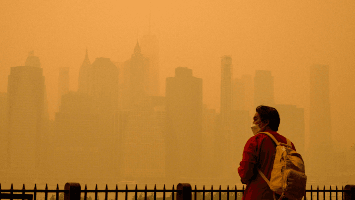 A person with dark hear wearing a red jacket and light-colored backpack stands in front of a New York City skyline shrouded in orange smoke from the wildfires