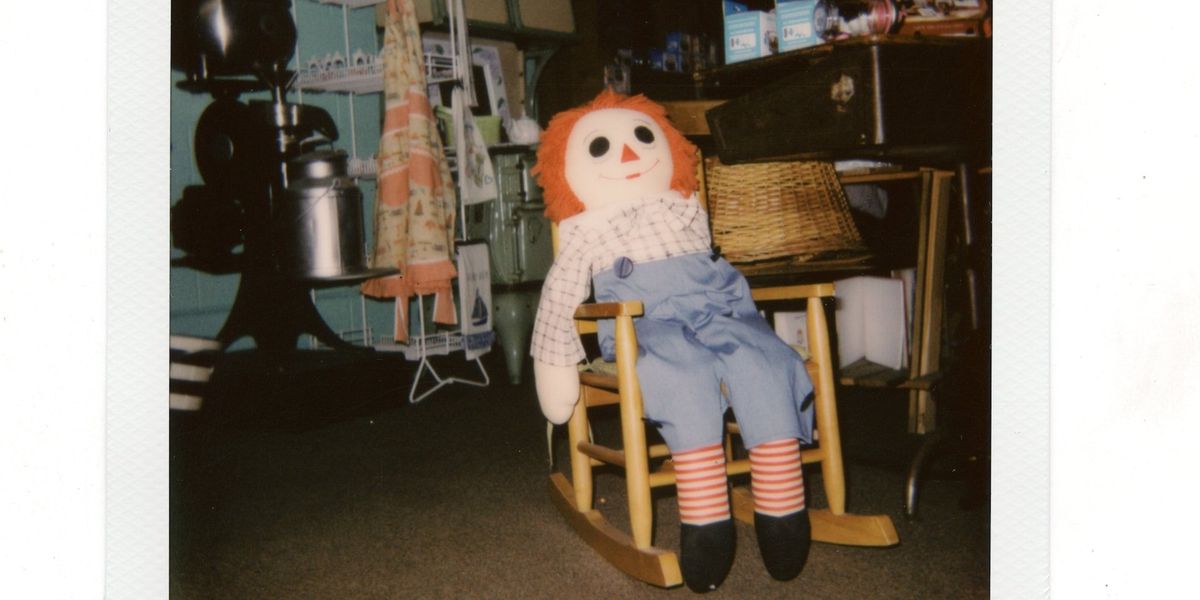 A polaroid of a creepy raggedy Anne doll sitting in a child's rocking chair surrounded by junk in a home