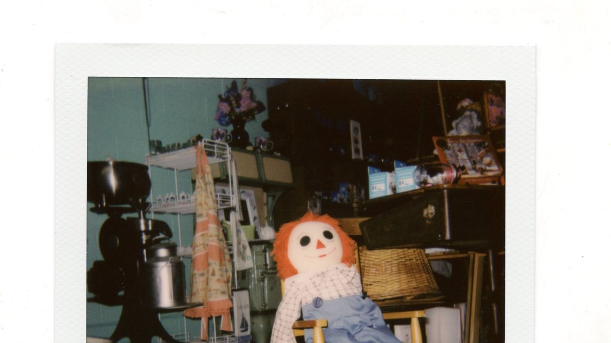 A polaroid of a creepy raggedy Anne doll sitting in a child's rocking chair surrounded by junk in a home