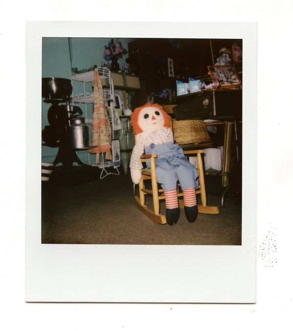A polaroid of a creepy raggedy Anne doll sitting in a child's rocking chair surrounded by junk in a home