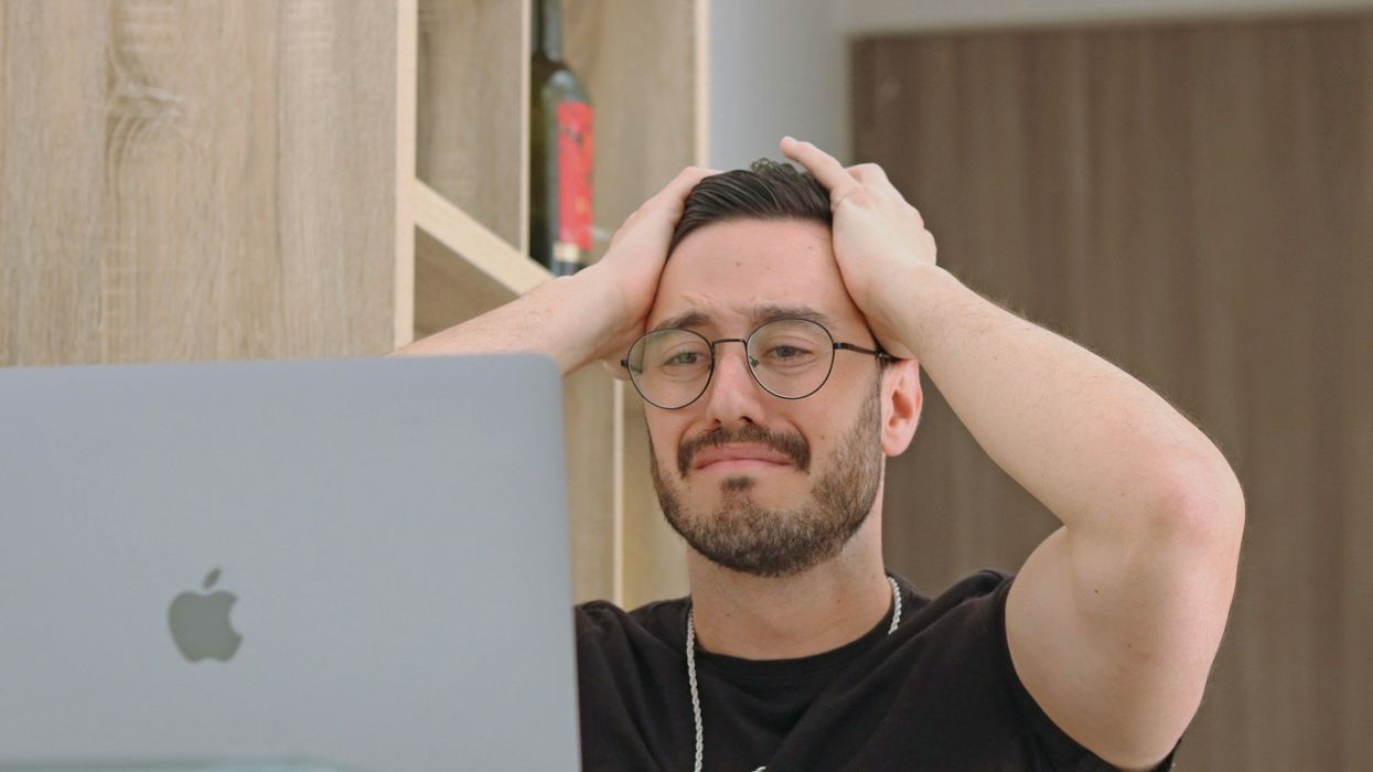 A sad young man receives unpleasant news on the computer. He has his hands on his and looks like he is about to cry.
