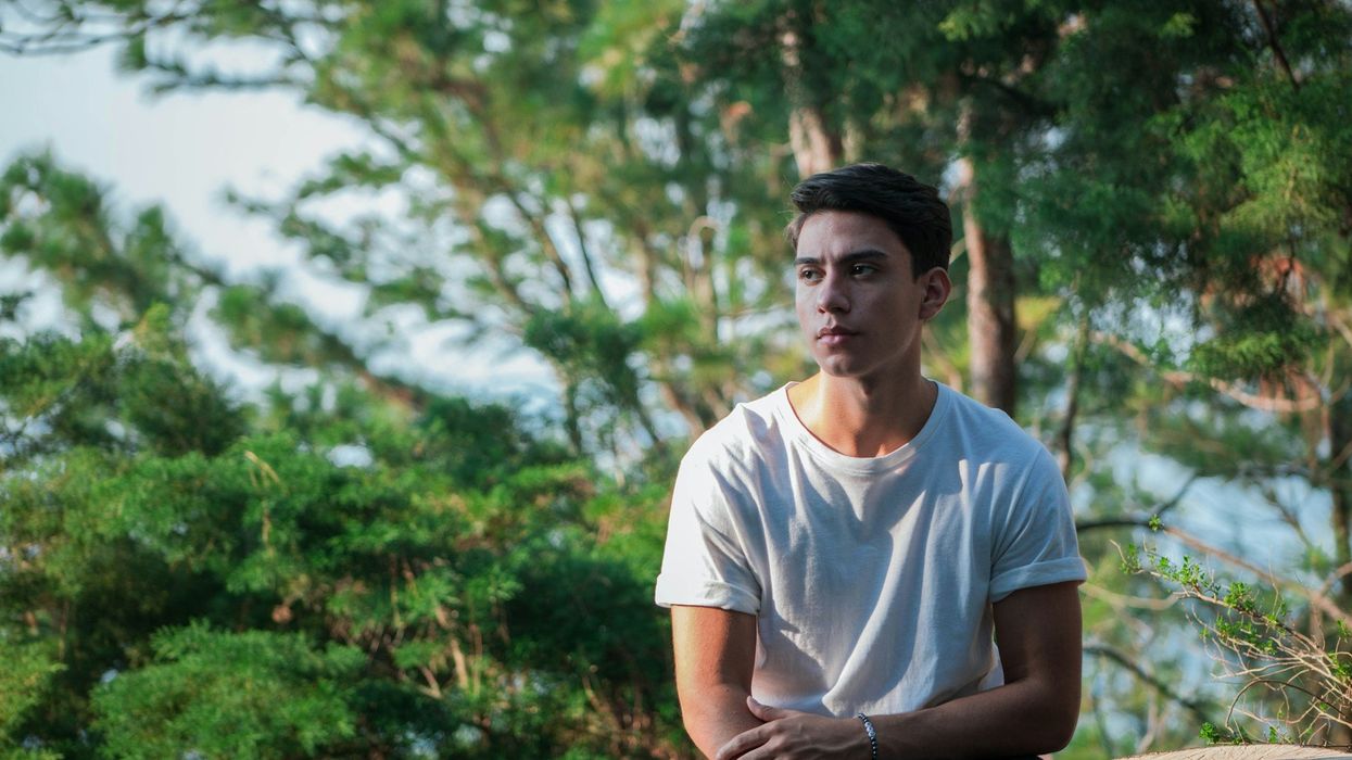 A shot of a young man sitting on a stump in the woods.