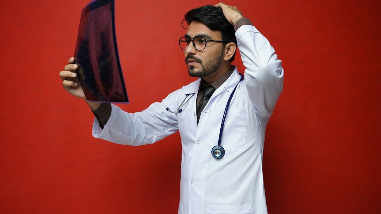 A slightly confused, young medical student, in a white lab coat looks at an e-ray. He stands against a red background with a stethoscope hanging around his neck.