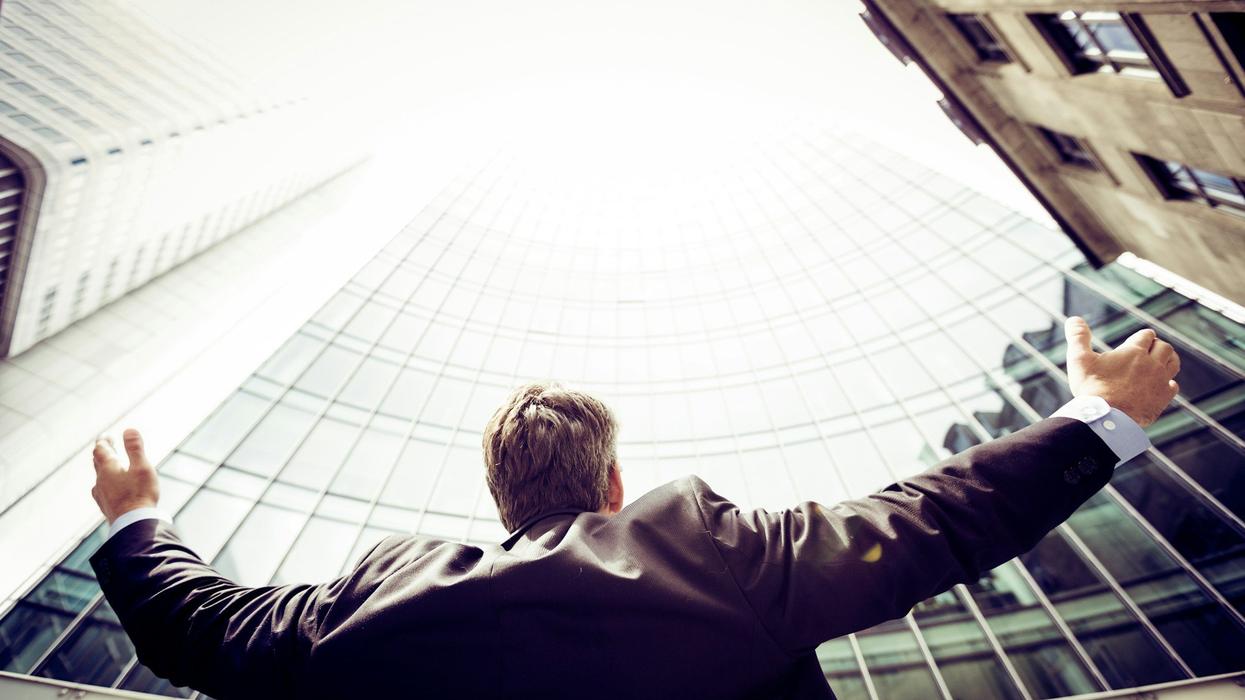 A wide back shot of a man, angled upward. He is in business attire and is staring in awe at a skyscraper.