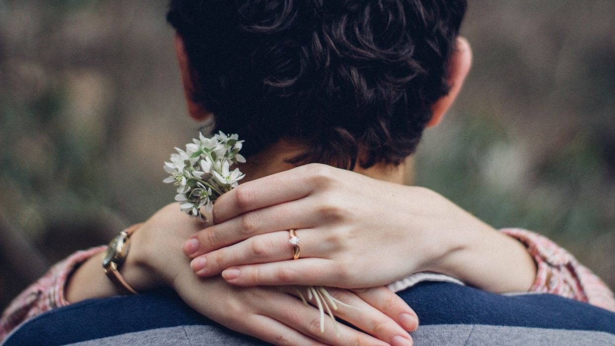 A woman has her hands around a man's neck. She wears an engagement ring and holds a small bouquet of white flowers.