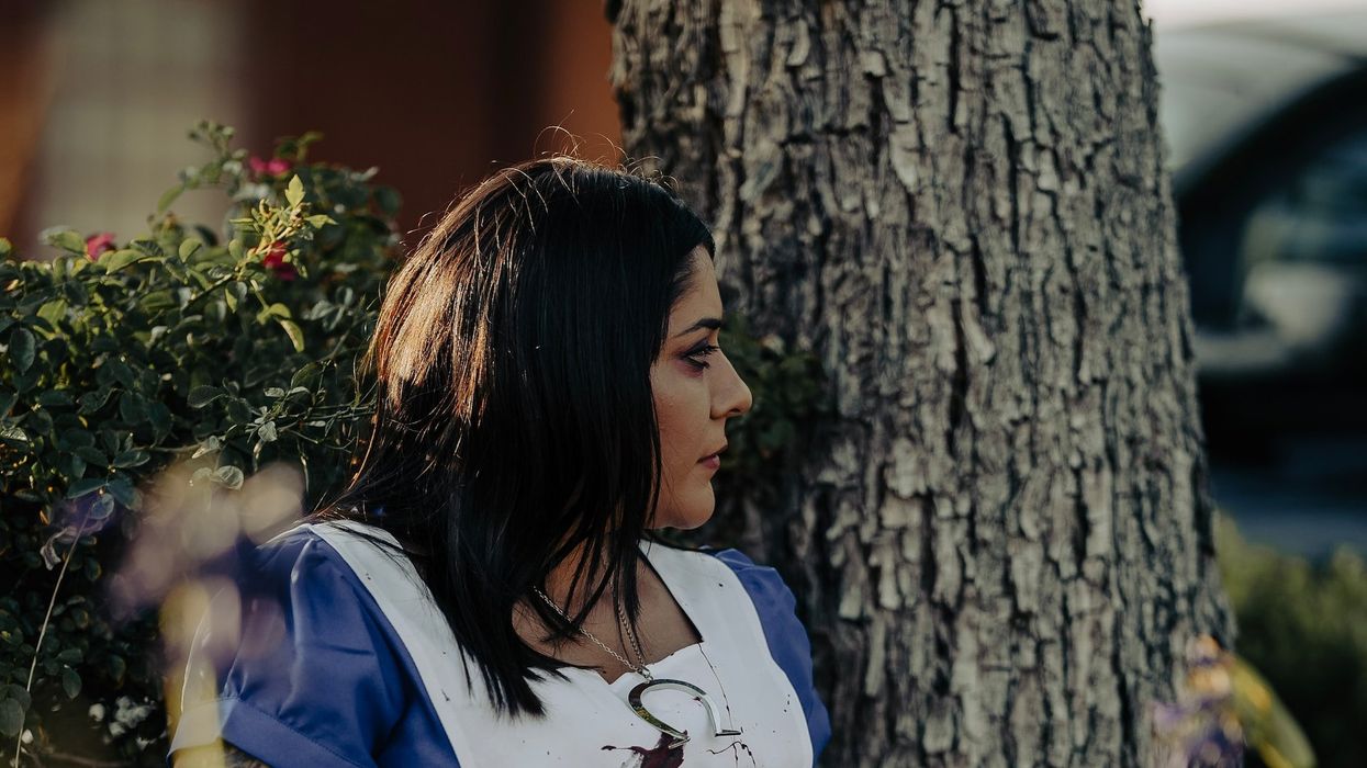 A woman hides behind a tree while she holds a knife and her front in splattered in blood