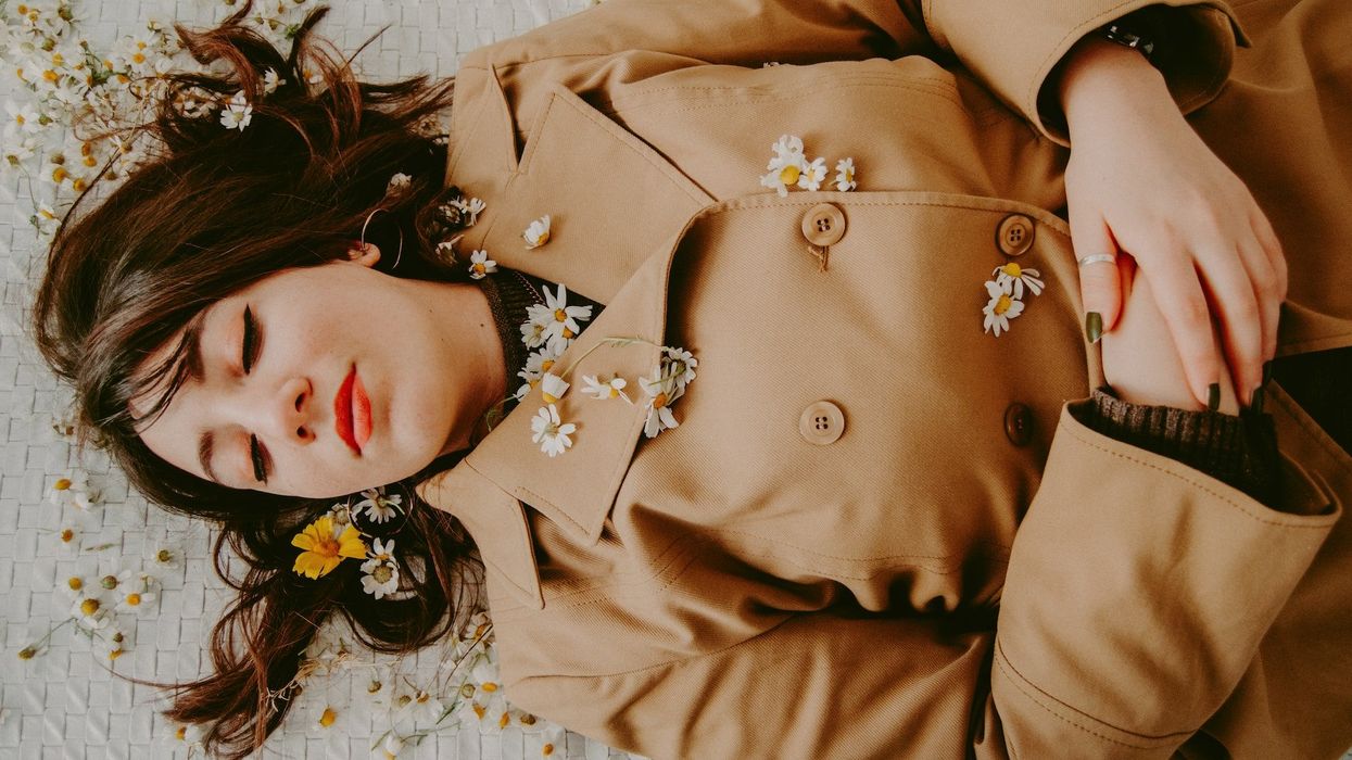 A woman lies on the ground, resting peacefully in a brown suede jacket, covered in small daisies