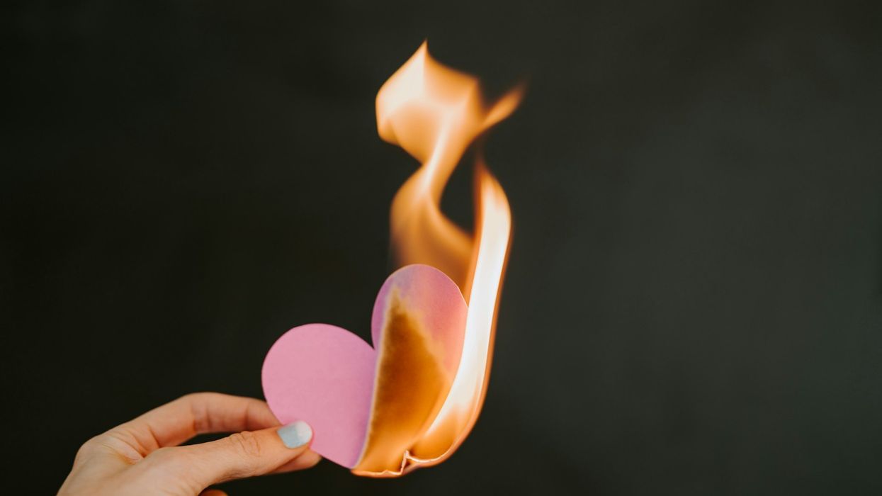 A woman's hand hold up a pink paper constructed heart that is on fire.