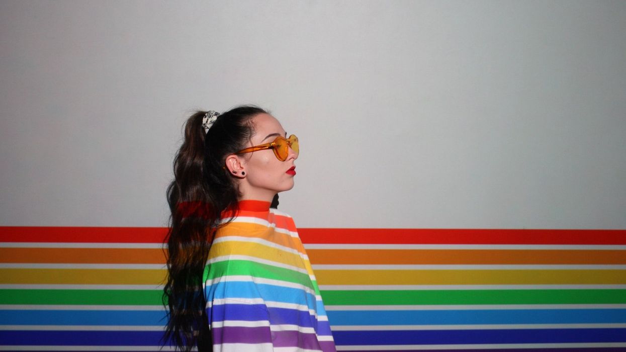 A woman wearing orange glasses stands against a white wall with the pride rainbow painted on it and her white jacket