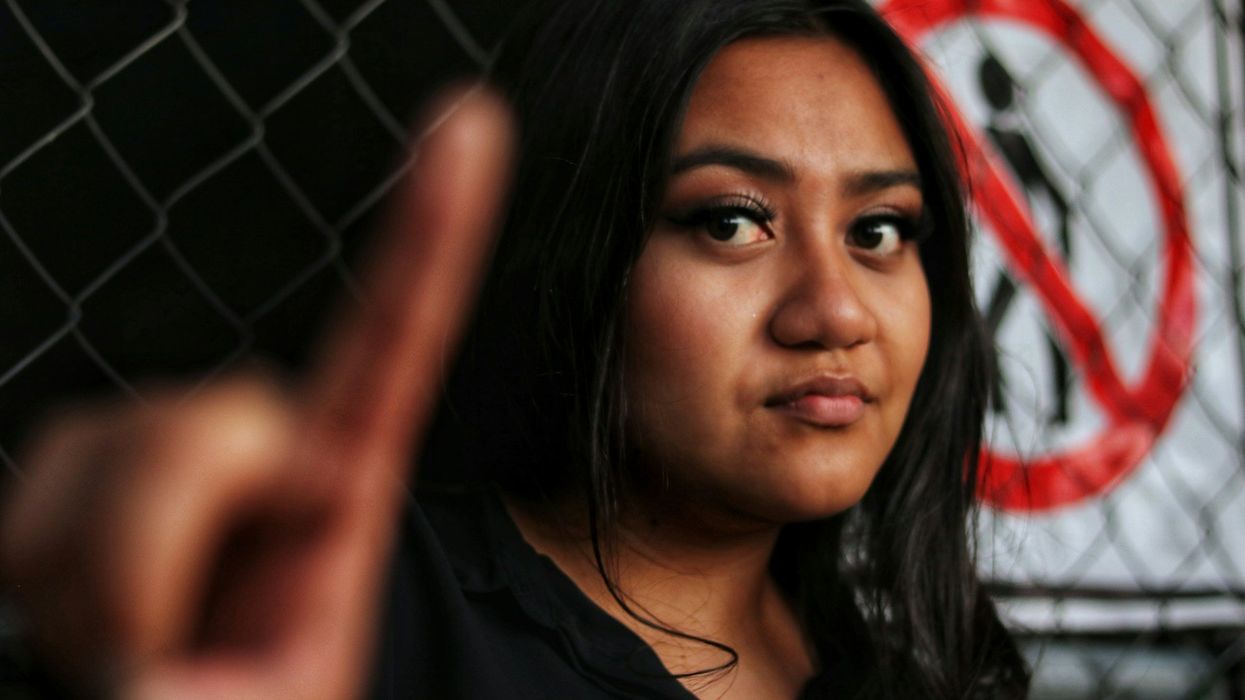 A young angered woman looks into the camera and sternly holds up her index finger. Behind her is a chainlink fence with a sign showing a stenciled person being barred.