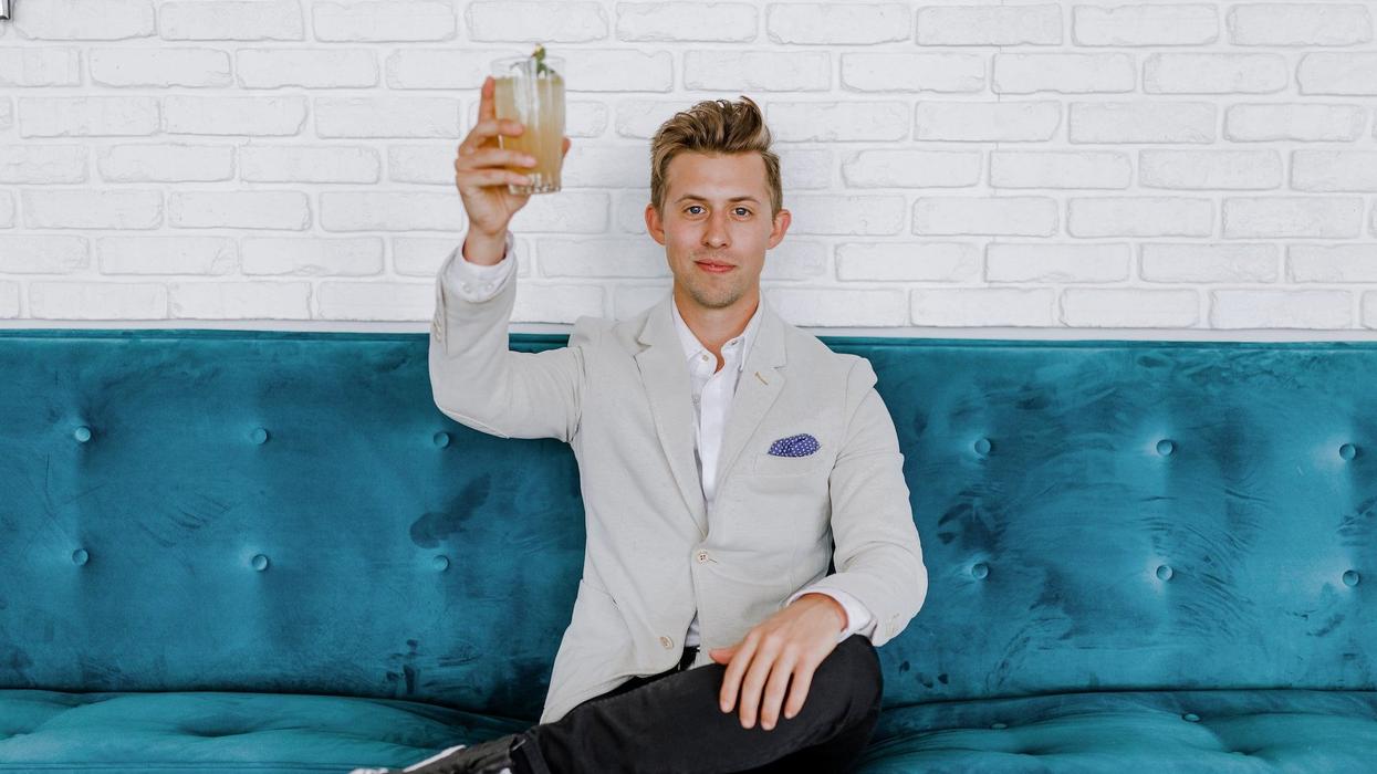 A young, attractive, fashionable man, sitting on a plush blue couch, gives a cheers