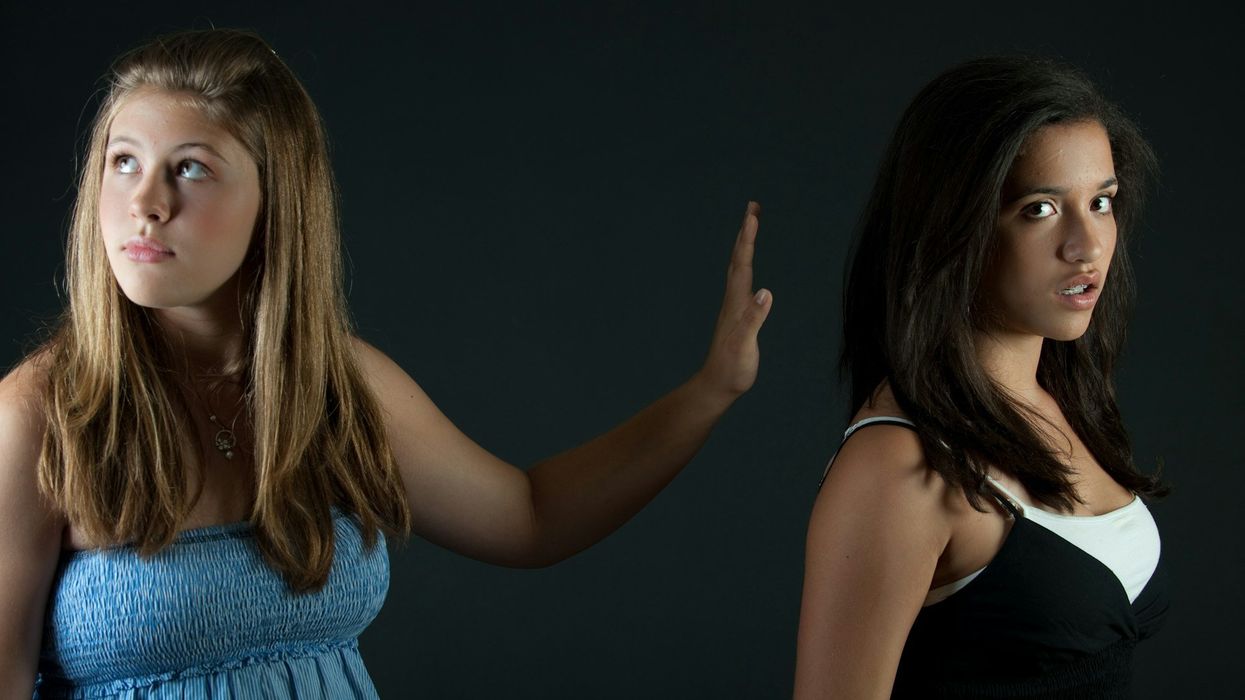 A young blonde girl in a light blue top rudely stretches out her hand to a young dark haired girl in a white and black top. The dark haired girl looks into the camera, the blonde girl looks up and away. They are standing in front of a dark background.
