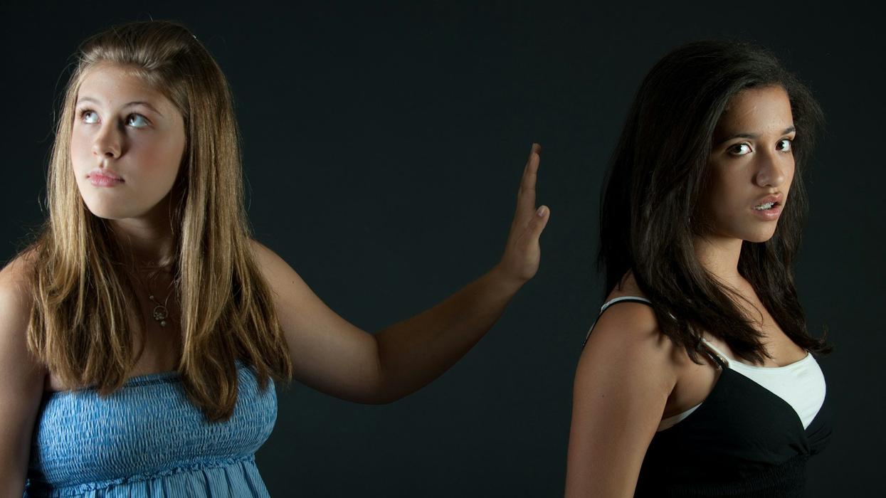 A young blonde girl in a light blue top rudely stretches out her hand to a young dark haired girl in a white and black top. The dark haired girl looks into the camera, the blonde girl looks up and away. They are standing in front of a dark background.