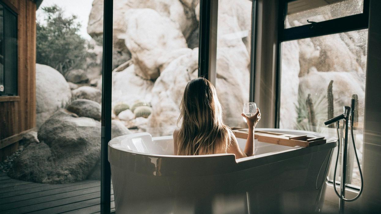 A young blonde woman baths in a luxury tub while holding a glass of wine, as she looks out the window. She has a view of majestic rocks.