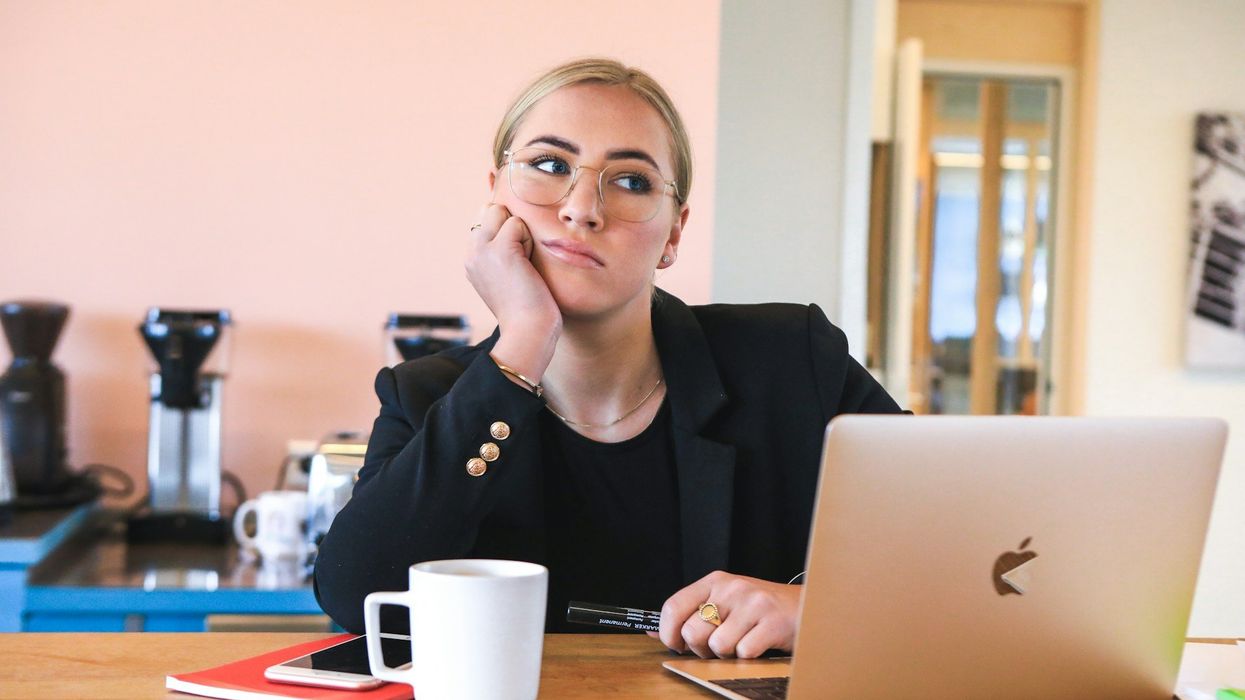 A young blonde woman in a black suit sits at her desk, her laptop is open and she is staring off in deep thought, she seems a bit perplexed.