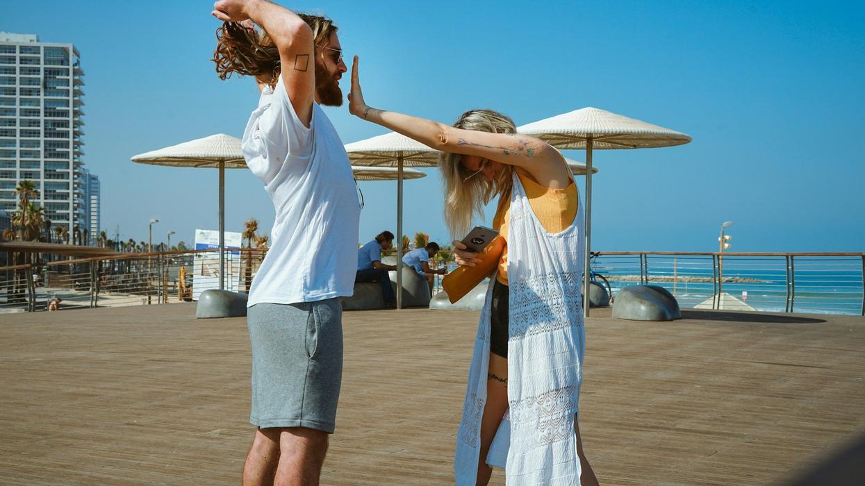 A young couple are on a boardwalk during a sunny day. The woman hangs her head and puts her hand's in her male partner's face to make him shut up.