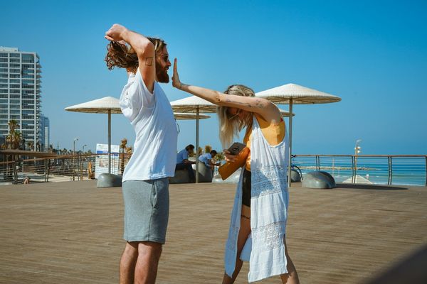 A young couple are on a boardwalk during a sunny day. The woman hangs her head and puts her hand's in her male partner's face to make him shut up.