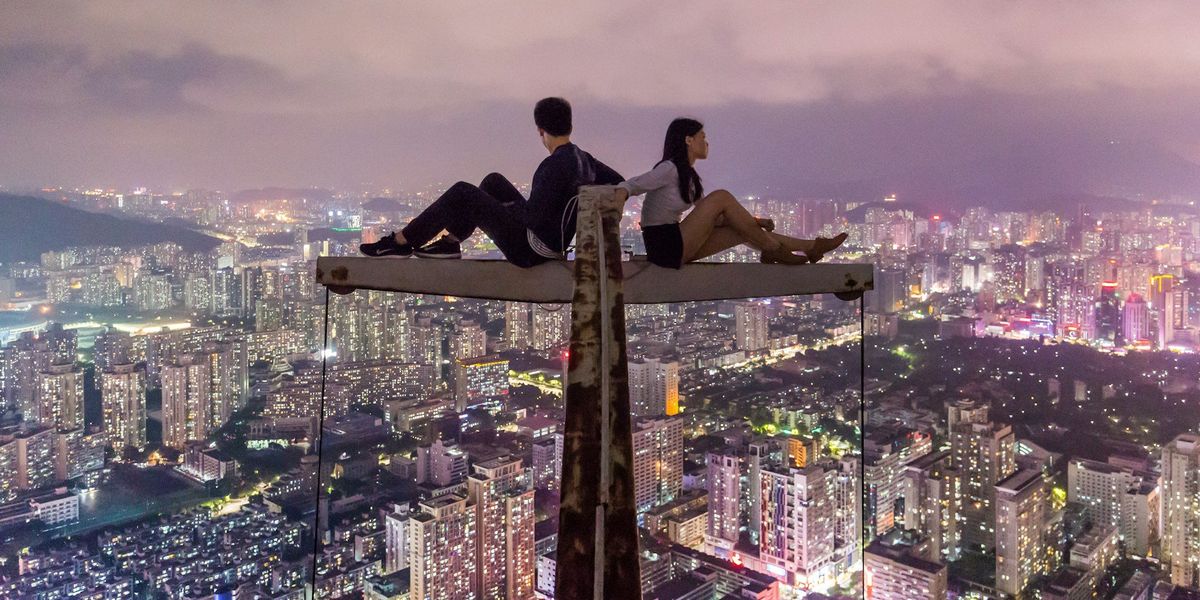 A young couple sit back to back on top of a crane overlooking the city at night.  