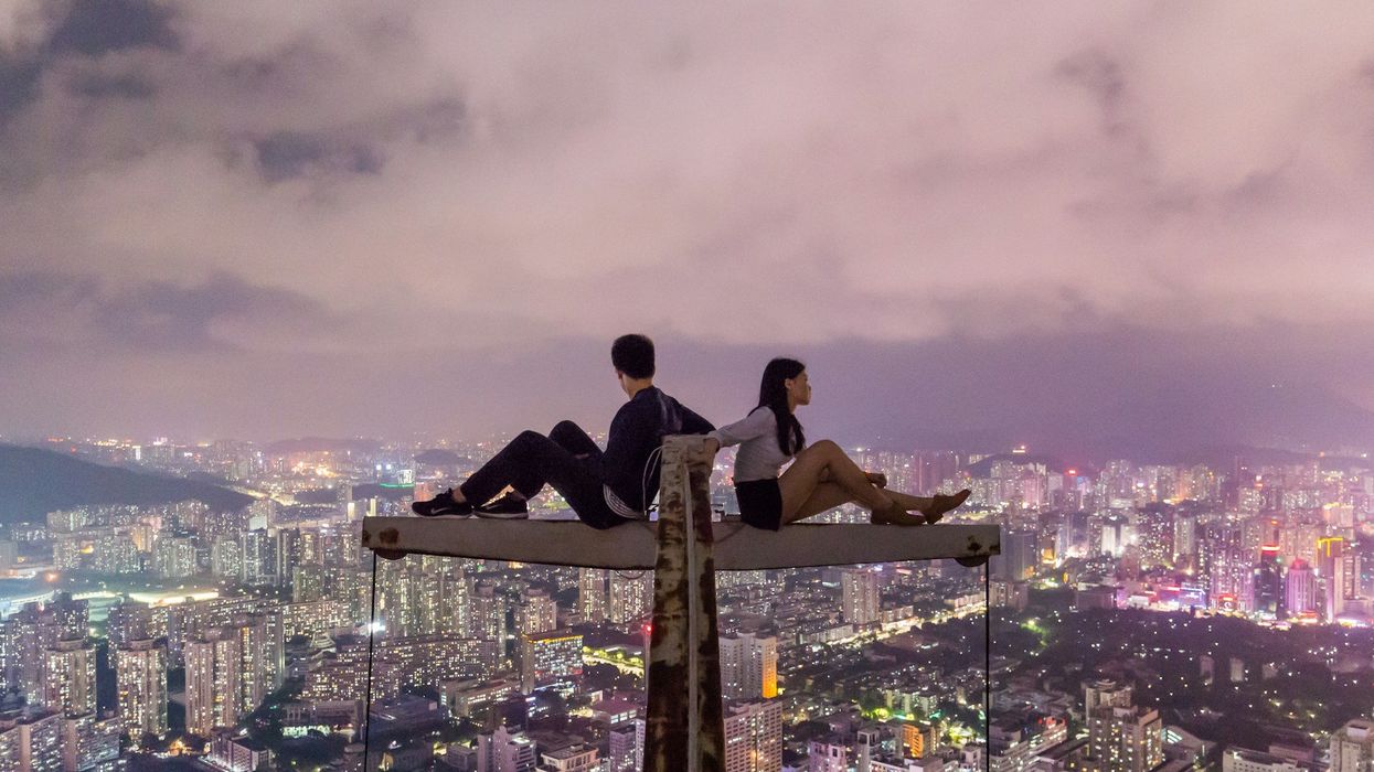 A young couple sit back to back on top of a crane overlooking the city at night.