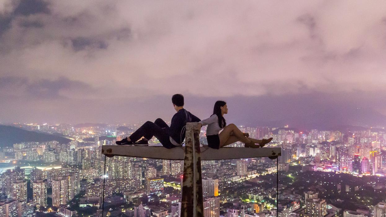 A young couple sit back to back on top of a crane overlooking the city at night.