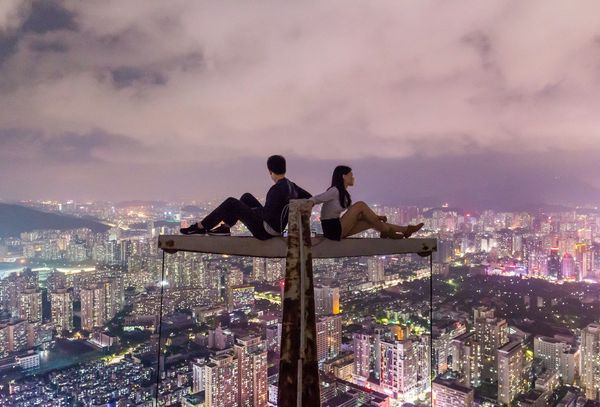 A young couple sit back to back on top of a crane overlooking the city at night.