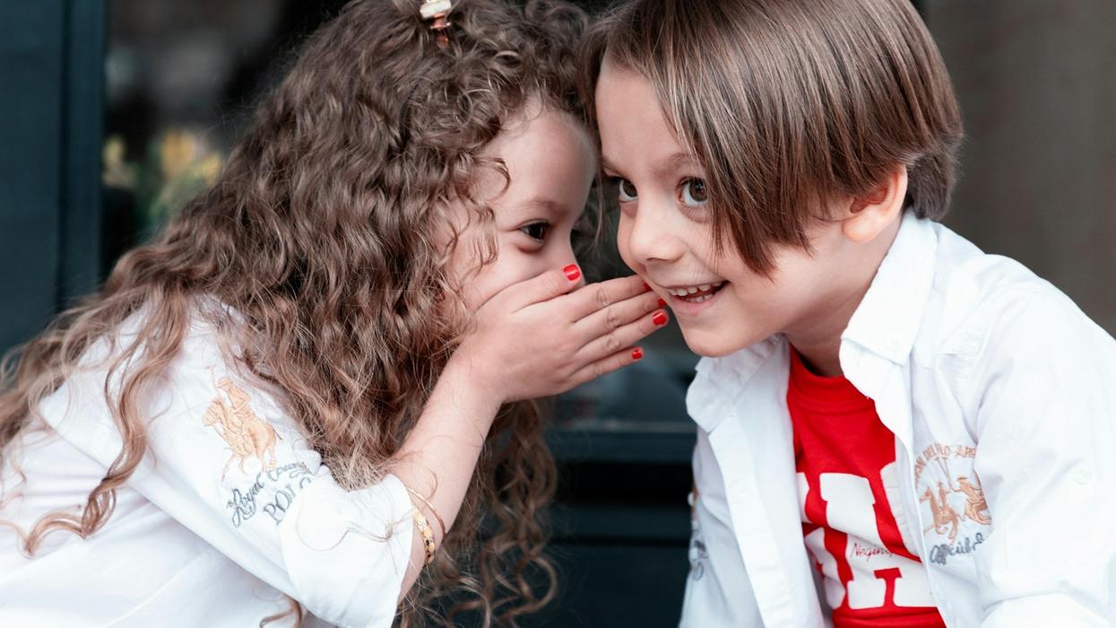 A young girl whispers secrets into her friend 's(a young man) ear. He sports a huge smile.