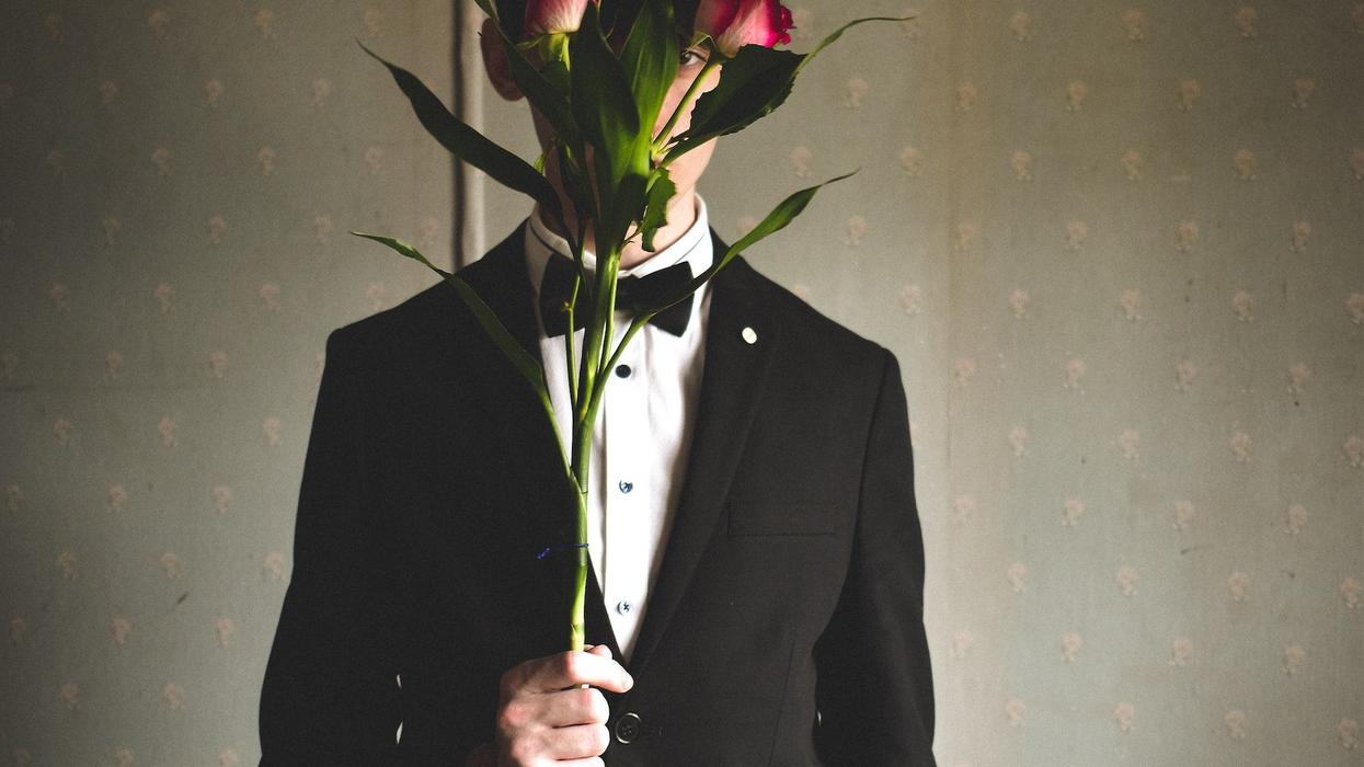 A young man in a tux, oddly holds two red roses in front of his face, only exposing one eye
