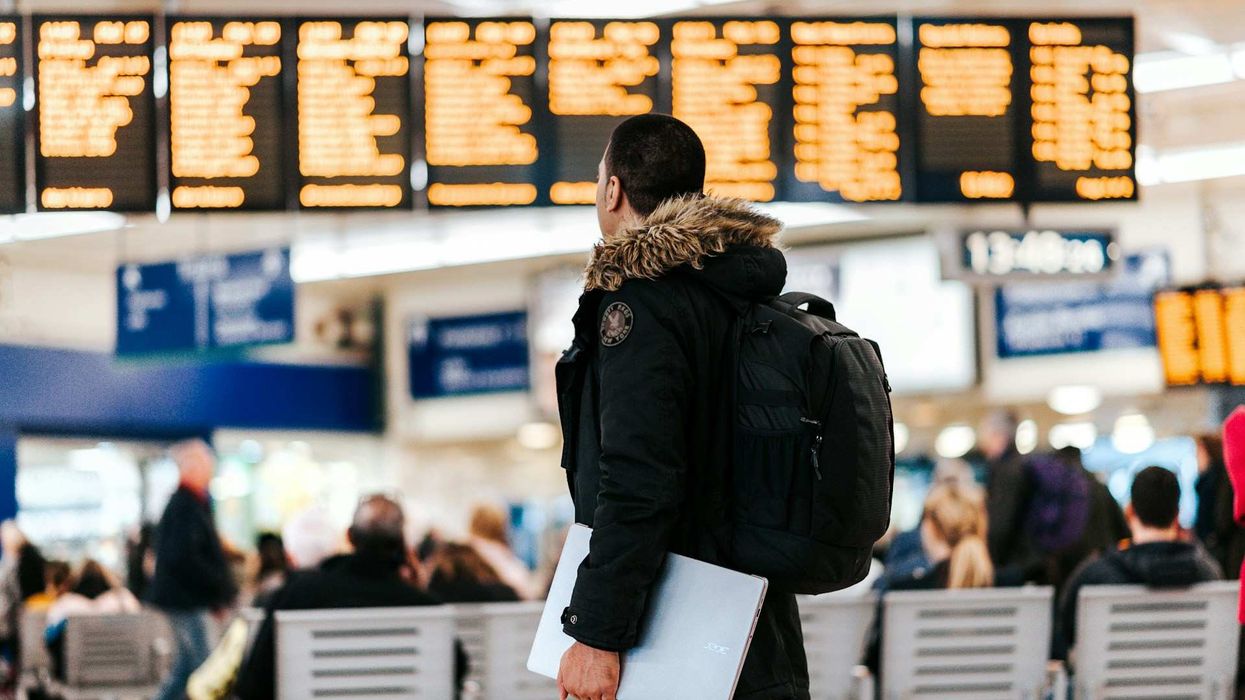 A young man in an airport wears a heavy winter coat and carries a laptop. He stares at the flight status board.